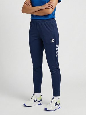 Persona in piedi con le braccia incrociate, indossa pantaloni della tuta Hummel blu scuro e scarpe sportive bianche con dettagli blu e verdi.