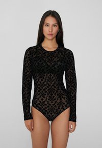 Czarny bodysuit z długim rękawem, wykonany z przezroczystej koronki z ozdobnym wzorem kwiatowym i podwyższonym dekoltem. Dopasowany kształt z wysokim krojem nogawki.