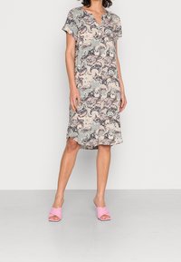 Mujer de pie con un vestido de patrón paisley, de longitud hasta la rodilla, con mangas cortas y sandalias de tacón abierto de color rosa, sobre un fondo claro.