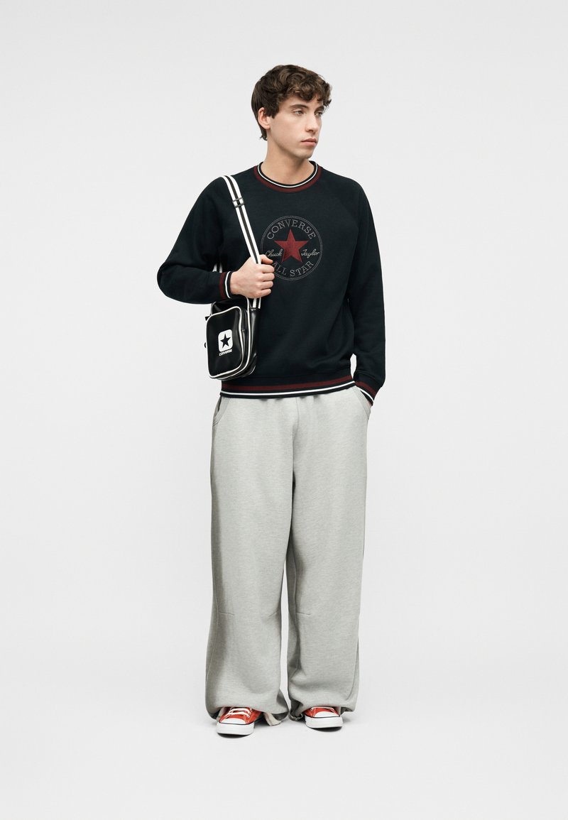 Young man wearing black Converse sweatshirt, gray wide pants, red sneakers, and carrying a black and white Converse crossbody bag.