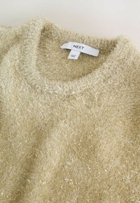 Pulover bej pufos cu guler rotund, având un fir metalic pe întreaga suprafață. Eticheta indică "NEXT", marcând brandul și țara de fabricație.