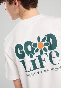 Valkoinen puuvillapaita, jossa on graafinen design, jossa lukee "Hyvä Elämä", kukka oranssilla keskustalla ja erilaisia luontoon liittyviä ikoneita.