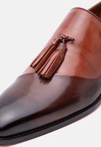 Δερμάτινο μοκασίνι δίχρωμο σε καφέ αποχρώσεις με διακοσμητικές φούντες στο επάνω μέρος, με στιλβωμένο, λείο φινίρισμα και ραμμένη άκρη στη σόλα.