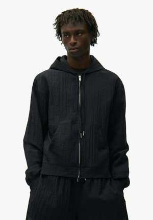 Giovane uomo con felpa nera con zip e trama, e pantaloni larghi abbinati, in piedi con le mani nelle tasche su sfondo bianco.