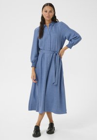Robe chemise bleue à manches longues en tissu texturé avec des boutons sur le devant, ceinture à nouer à la taille et manches bouffantes, assortie à des chaussures noires.