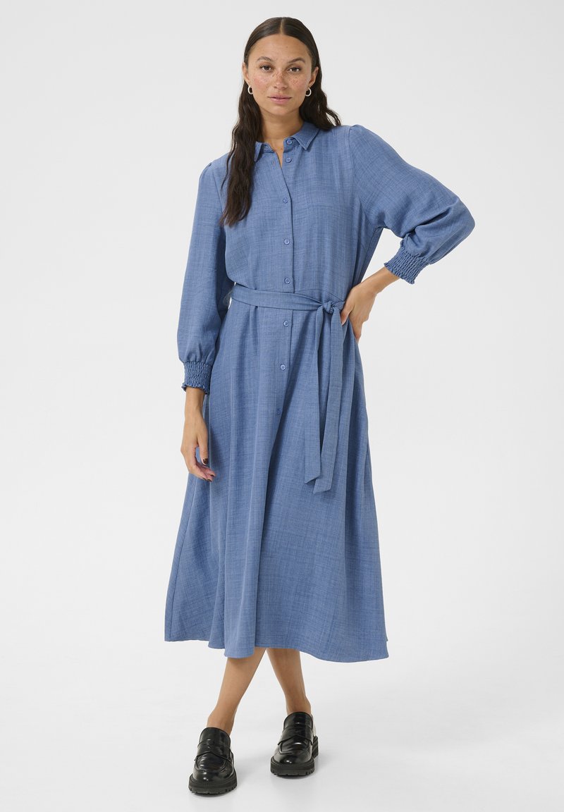 Robe chemise bleue à manches longues en tissu texturé avec des boutons sur le devant, ceinture à nouer à la taille et manches bouffantes, assortie à des chaussures noires.