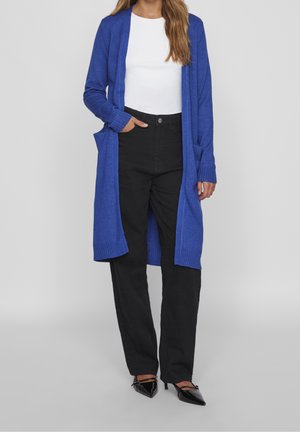 Femme portant un long cardigan bleu, un haut blanc, un pantalon noir taille haute et des talons pointus noirs, debout devant un fond clair uni.