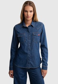 Camisa de mezclilla en azul oscuro con dobles bolsillos en el pecho, costuras naranjas y diseño de yugo puntiagudo. Mangas largas con puños de botón.