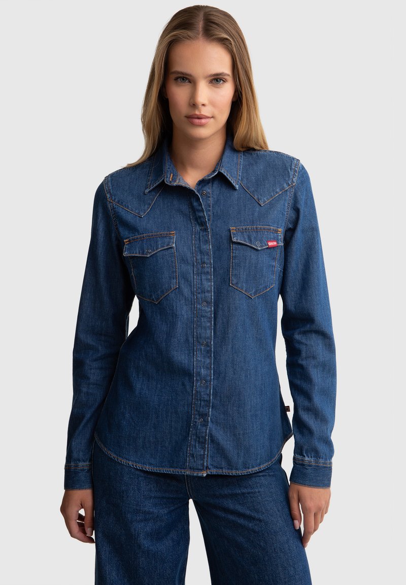 Camisa de mezclilla en azul oscuro con dobles bolsillos en el pecho, costuras naranjas y diseño de yugo puntiagudo. Mangas largas con puños de botón.