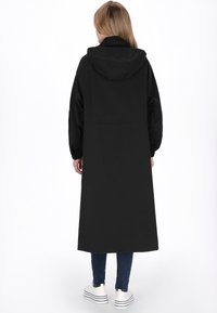 Fekete hosszú kabát kapucnival, egyenes sziluettel, elasztikus mandzsettákkal és sima textúrával, kék farmernadrág és fehér cipő felett viselve.