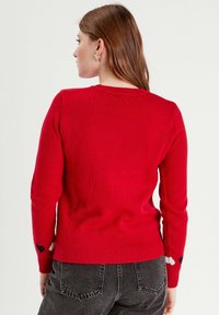 Pull en maille rouge avec des manches longues, ourlet et poignets côtelés. Présente des motifs de cœurs sur les manches, une texture lisse et une encolure ras du cou classique.