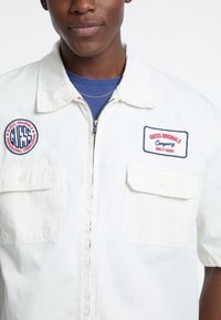 Giacca bianca con zip, dotata di due tasche sul petto, toppe ricamate e maniche corte. Realizzata in un tessuto leggero.