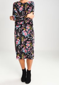 Abito nero a maniche lunghe con un motivo floreale colorato, caratterizzato da fiori rosa, blu e gialli; design aderente, lunghezza fino al ginocchio, indossato con stivaletti neri. 