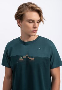 Katoenen T-shirt in diep teal, met een bedrukte afbeelding van boten en heuvels. Korte mouwen, ronde hals en subtiele textuur.