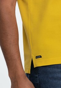 Gul tekstureret polo shirt med sideskår og et sort "ombre" label i kanten, parret med mørke denim jeans.