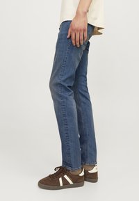 Jeans de mezclilla azul con un corte recto, diseño de cinco bolsillos y un lavado medio. Combinados con zapatillas de ante marrón con rayas blancas.