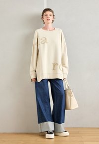 Beige oversized sweatshirt met lichtbruine lettering, gecombineerd met denim jeans met wide-leg en rafelige uiteinden, en zwarte platform sneakers.