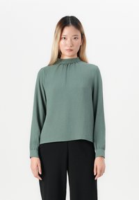 Kvinne med langt, rett hår som har på seg en grønn bluse med lange ermer og rynket høy hals, sammen med svarte bukser, står mot en hvit bakgrunn.