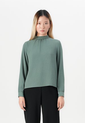 Woman with long straight hair wearing a green long-sleeve blouse with a gathered high neckline and black pants, standing against white background.