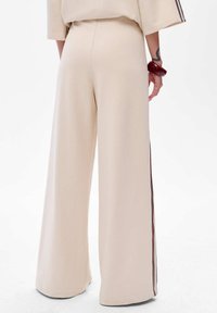 Beige wijdvallende broek van zachte stof, met een gladde textuur en contrasterende streep aan de zijkanten. Geen zichtbare sluitingen.