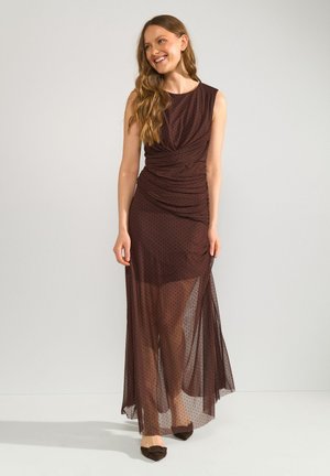 Femme aux longs cheveux ondulés portant une robe maxi sans manches, marron foncé, transparente à pois et des ballerines pointues foncées, souriante et regardant sur le côté.
