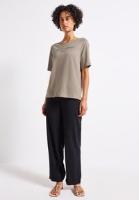 Khaki t-shirt med korta ärmar som har svart text, i kombination med lösa svarta byxor och beige sandaler mot en neutral bakgrund.