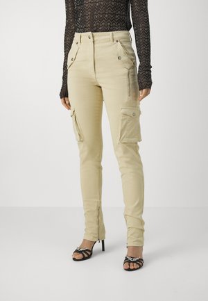 Beige cargobroek met meerdere voorkruimelzakken, slim fit en ritsen bij de enkels, gecombineerd met zwarte hoge hakken.