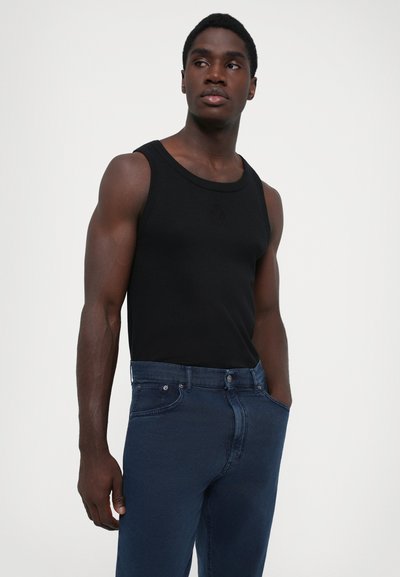 Muscular man wearing a black sleeveless top and blue jeans, standing with hands in pockets and looking to the side against a plain white background.