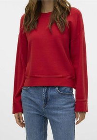 Sweat-shirt rouge à coupe décontractée, encolure ronde et épaules tombantes. Poignets côtelés ; porté avec un jean bleu taille haute.