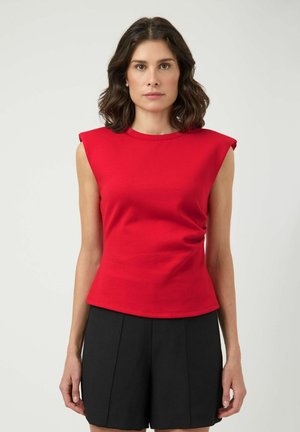 Vrouw met donker schouderlang haar, gekleed in een mouwloze rode top met gewatteerde schouders en een zwarte short met hoge taille, staand tegen een effen achtergrond.