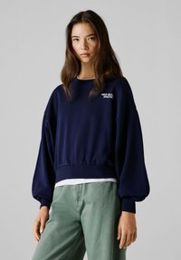 Sudadera corta azul marino con bajo acanalado, cuello redondo y mangas abullonadas. Presenta texto blanco "RED BULL" en el frente.