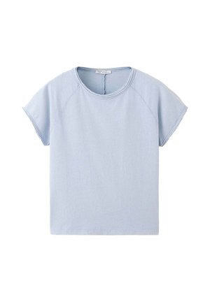 Jonge vrouw staat tegen een effen achtergrond, draagt een lichtblauw topje met korte mouwen over een gestreept overhemd, ruime donkere jeans en zwarte schoenen.