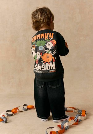 Barn med kort hår iført en sort Halloween-sweatshirt med tegneseriefigurer, stående omgivet af orange, hvide og sorte papkæde dekorationer.