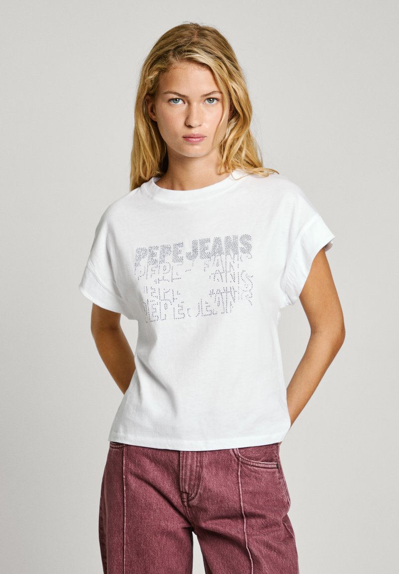 T-shirt en coton blanc avec des manches roulées et un design de texte scintillant. Associé à un pantalon en denim rose, avec une coupe décontractée.