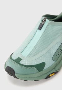 Chaussure slip-on vert clair avec fermeture éclair, tige en maille texturée, semelle en caoutchouc et semelle extérieure noire avec la marque Vibram.