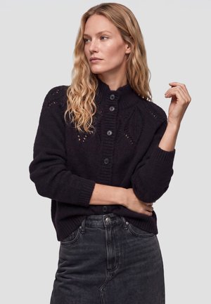 Kvinde med bølget blondt hår iført en sort knap-trøje og mørkt denimskørt, poserer med den ene arm krydset og den anden løftet nær skulderen.