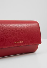 Pochette Emporio Armani en cuir texturé rouge avec logo doré sur le devant, présentée sur un fond clair uni.