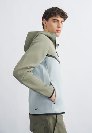 Mladý muž v zelenej a svetlosivej mikine na zips so vstavanou kapucňou, so zastrčenou rukou v vrecku, stojaci z profilu proti jednoliatemu bielemu pozadiu.