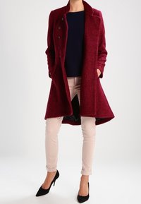 Bordeaux wollen jas met een geknipte kraag, zes knopen, twee zijzakken en een getailleerde silhoeet, gecombineerd met lichte broek en zwarte hakken.