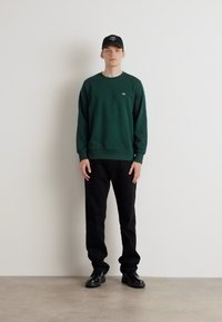 Groene sweatshirt met geribde manchetten, zwarte broek en zwarte schoenen. Model draagt een zwarte pet. Effen achtergrond, volledig lichaamsperspectief.