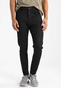 Černé džínové kalhoty slim fit, se pěti kapsami, zapínáním na knoflík a plochým předním dílem. Nosené se šedými sportovními botami.