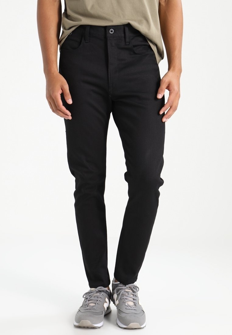 Černé džínové kalhoty slim fit, se pěti kapsami, zapínáním na knoflík a plochým předním dílem. Nosené se šedými sportovními botami.
