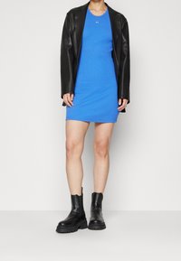 Kobieta w niebieskiej, bezrękawnikowej mini sukience, czarnej skórzanej kurtce i czarnych, masywnych botkach na kostkę stoi na jednolitym tle.