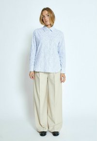 Camicia azzurra chiara a maniche lunghe con motivo di diamanti a rilievo, colletto a punta e orlo irregolare, abbinata a pantaloni larghi crema e scarpe nere.