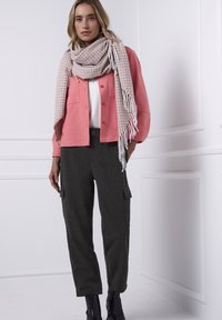 Lichtroze jasje met knopen over een wit shirt, gecombineerd met donkere geruite cargobroek en een sjaal met patroon en franjes. Zwarte enkellaarzen.