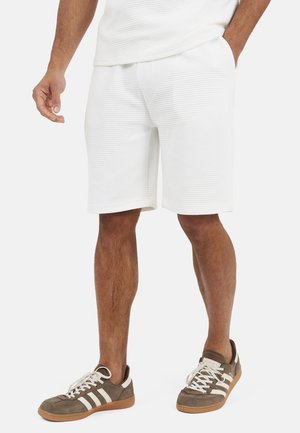 Man draagt witte textuur korte broek en bruine Adidas sneakers met witte strepen, staand met één hand in de zak.