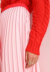 Pull en laine rouge tricoté avec un motif texturé et des manches longues, associé à une jupe plissée rose présentant de fines lignes verticales et une taille élastique.