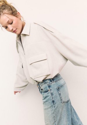 Femme aux cheveux blonds portant une veste oversize beige clair avec de grandes poches frontales et un jean en denim bleu taille haute.
