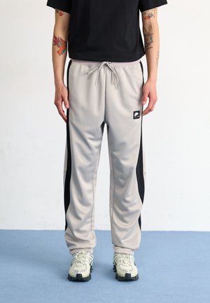 Persoon die lichtgrijze joggingbroek draagt met zwarte zijpanelen, witte sneakers en een zwart T-shirt met korte mouwen, staand op een blauwe vloer.