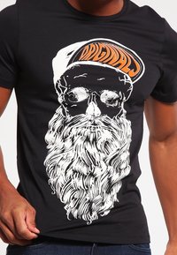 La camiseta negra presenta un gran gráfico blanco de una cara con barba y gafas de sol, con texturas detalladas en el cabello; "ORIGINAL" en naranja en una gorra.